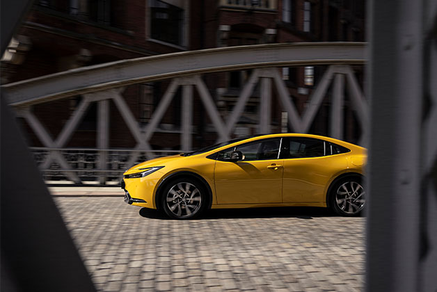 Na zdjęciu widoczny jest żółty samochód Toyota Prius jadący po ulicy z kostki brukowej. W tle znajduje się most o stalowej konstrukcji. Zdjęcie zostało zrobione pod kątem, z widocznymi elementami ramy po obu stronach kadru, co nadaje mu dynamiczny charakter.