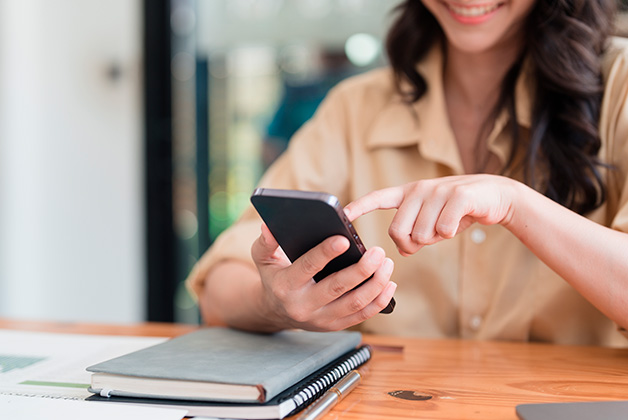 Zdjęcie przedstawia uśmiechniętą kobietę z długimi, ciemnymi włosami, siedzącą przy drewnianym biurku, trzymająca w dłoniach telefon. Obok na biurku leżą notesy oraz długopis. Tło zdjęcia jest rozmyte.