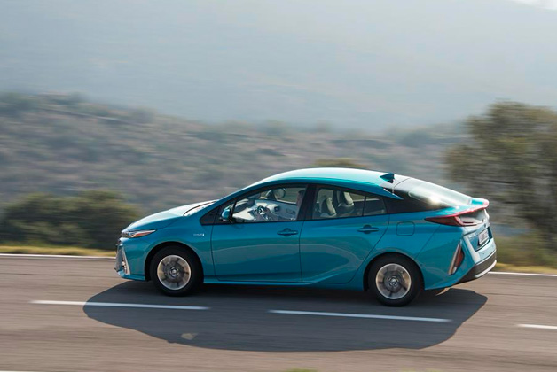 Zdjęcie przedstawia Toyotę Prius, w kolorze niebieskim, jadącą po asfaltowej drodze.  Samochód jest w ruchu, co sugeruje rozmyte tło, na którym widać porośnięte roślinnością doliny. 