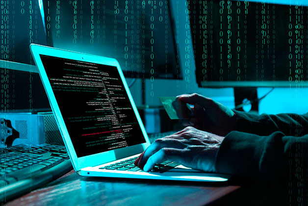 Ręce osoby pracującej przy laptopie, na ekranie kod komputerowy, w tle monitory – jedna dłoń trzyma kartę płatniczą; ilustracja zagrożeń związanych z bezpieczeństwem danych.