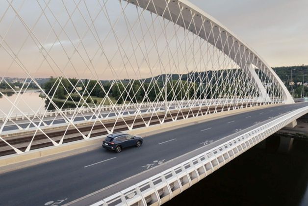 Na zdjęciu widoczna ciemna Toyota Corolla, jadąca przez szeroki, biały most. Wzdłuż jezdni widoczne są oznaczenia ścieżki rowerowej. W tle rozpościera się zielony, pagórkowaty krajobraz z widocznymi budynkami po drugiej stronie rzeki.