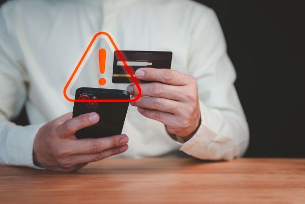 Zdjęcie przedstawia człowieka trzymającego w dłoni telefon komórkowy oraz kartę bankową. Nad telefonem i kartą unosi się duży, czerwony trójkąt z wykrzyknikiem, symbolizujący ostrzeżenie lub niebezpieczeństwo. Kompozycja obrazu sugeruje zagrożenie związane z oszustwami finansowymi w sieci, phishingiem lub niebezpiecznymi transakcjami internetowymi.