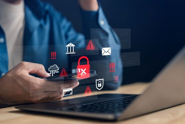 Zdjęcie przedstawia osobę korzystającą z bankowości mobilnej na telefonie. Nad dłonią trzymającą smartfon unosi się cyfrowy obrazek otwartej kłódki z iksem. Wokół telefonu i laptopa, symbolizującego urządzenie do bankowości internetowej, widoczne są różne ikony i piktogramy, takie jak wykrzykniki, bank, chmura, tarcza, koperta, które są częściowo przesłonięte przez czerwone ostrzeżenia i przekreślenia. Wszystko to symbolizuje cyberzagrożenia w bankowości mobilnej.