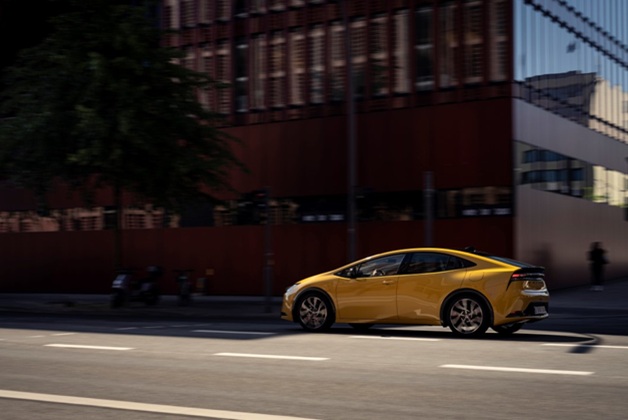 Toyota Prius w kolorze żołtym poruszająca się po miejskiej ulicy w słoneczny dzień. W tle widoczny jest duży, nowoczesny budynek z licznymi oknami, odbijający światło.