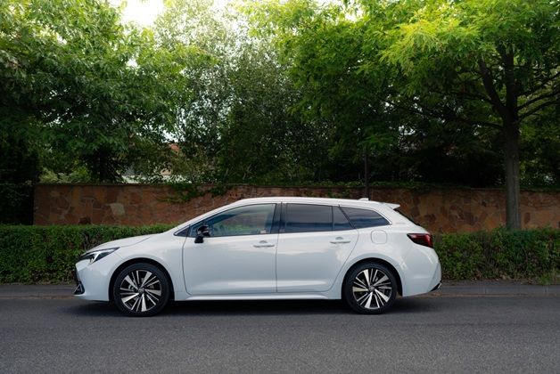 Biała Corolla TS Kombi zaparkowana przy ulicy, otoczona zielenią i drzewami, w tle kamienny mur i krzewy