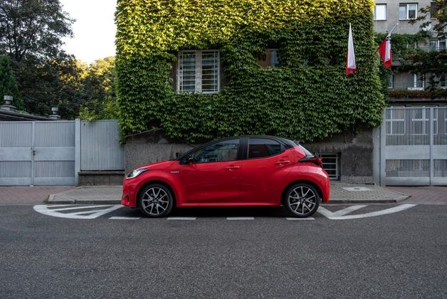 Czerwony hatchback - Toyota Yaris - zaparkowany na ulicy, na tle budynku całkowicie pokrytego bluszczem, z widoczną polską flagą.