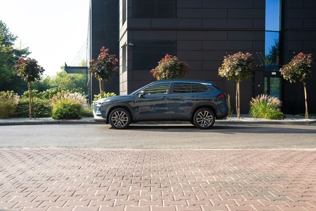 Szara Corolla Cross zaparkowana bokiem przy ulicy obok nowoczesnego budynku z ciemną elewacją i małymi drzewkami ozdobnymi.