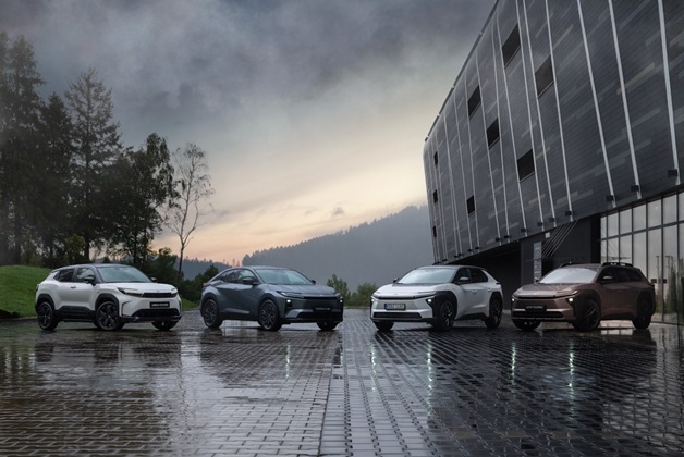Cztery elektryczne SUV-y Toyota bZ4X Touring w różnych kolorach (od lewej: biały, szary, biały i brązowy) zaparkowane w rzędzie na mokrej, wybrukowanej powierzchni obok nowoczesnego budynku, z zielonymi wzgórzami i zachmurzonym niebem w tle.
