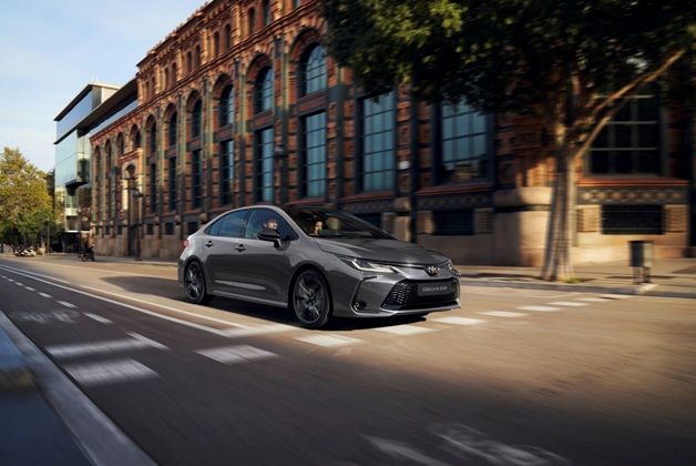 Zdjęcie przedstawia ciemnoszary samochód Toyota Corolla Sedan jadący ulicą obok dużego, starego budynku z cegły z wieloma oknami. W aucie widoczne są dwie osoby.