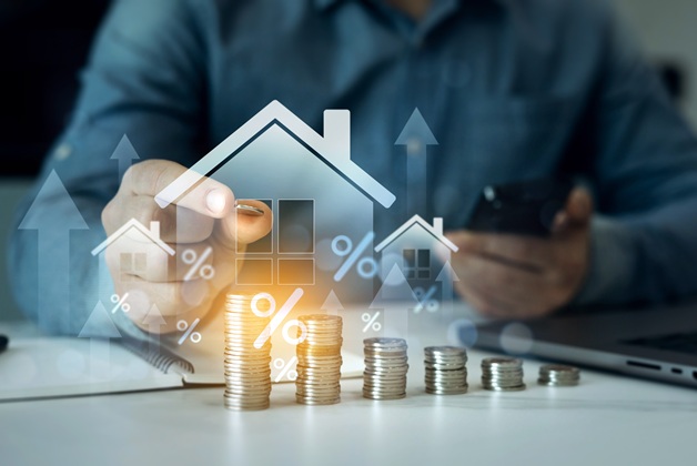 Zdjęcie przedstawia symboliczną wizualizację wzrostu stóp procentowych i cen nieruchomości z ikonami domów i stosami monet.