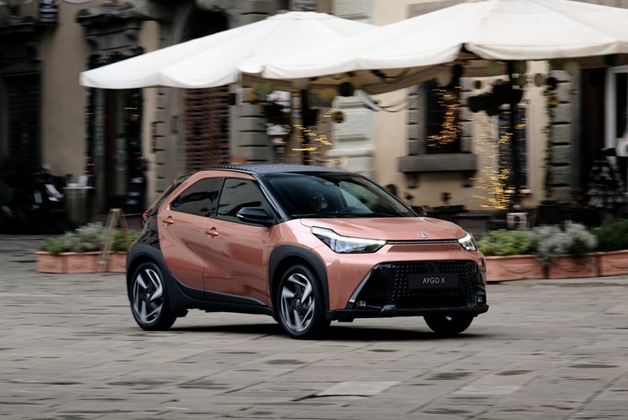 Zdjęcie przedstawia nowoczesny, miedziano-czarny miejski crossover Toyota Aygo X jadący po brukowanym placu na tle kawiarnianych parasoli i zabytkowych kamienic. 