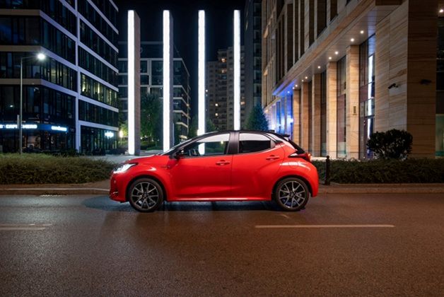 Zdjęcie Toyota Yaris w czerwonym kolorze. Samochód jest zaparkowany bokiem na miejskiej ulicy. W tle widoczna jest nowoczesna architektura z podświetlonymi pionowymi filarami, które tworzą efektowny kontrast z ciemnym, wieczornym otoczeniem.
