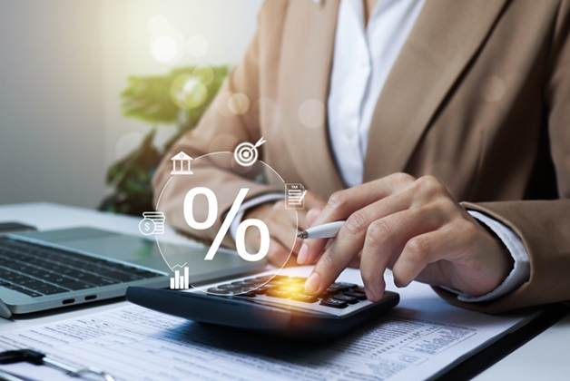 Osoba w beżowej marynarce korzysta z kalkulatora przy laptopie. Nad urządzeniem widnieje cyfrowa ikona procenta oraz symbole finansowe: bank, wykresy i dokumenty.