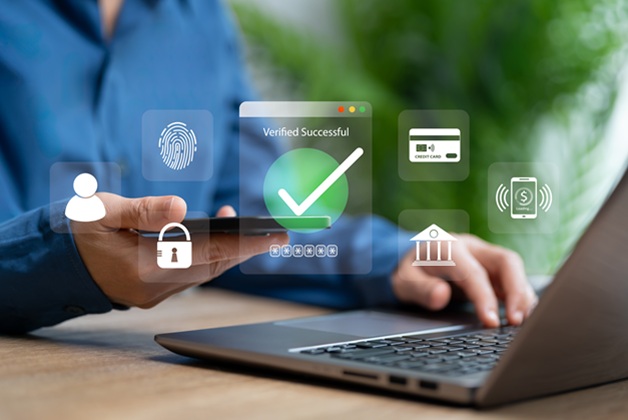  Osoba korzystająca jednocześnie z laptopa i smartfona, nad którymi wyświetlają się cyfrowe ikony zabezpieczeń: kłódka, odcisk palca, karta płatnicza i budynek banku.