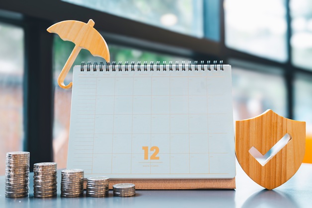 Zdjęcie przedstawiające biały kalendarz z liczbą 12, obok którego znajdują się rosnące stosy monet, mały żółty parasol oraz drewniana tarcza z symbolem „v”. Całość symbolizuje planowanie finansowe, oszczędzanie i ochronę kapitału.