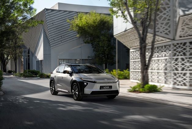 Nowoczesny, elektryczny SUV Toyota bZ4X w kolorze srebrnym, poruszający się po asfaltowej drodze obok białego budynku z dekoracyjną fasadą.