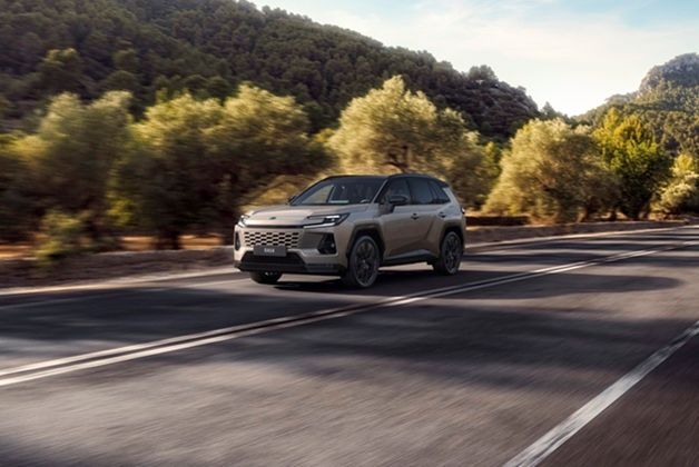 Nowa Toyota RAV4 w kolorze jasnobeżowym, jadąca dynamicznie po asfaltowej drodze wśród zielonych wzgórz i drzew oliwnych. Samochód prezentuje nowoczesny, kanciasty design z charakterystycznymi reflektorami LED i wyrazistą osłoną chłodnicy.