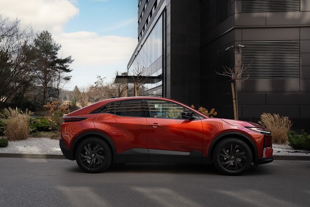 Nowy elektryczny SUV Toyota C-HR+ w kolorze pomarańczowym, widok z boku na tle ciemnej elewacji biurowca.