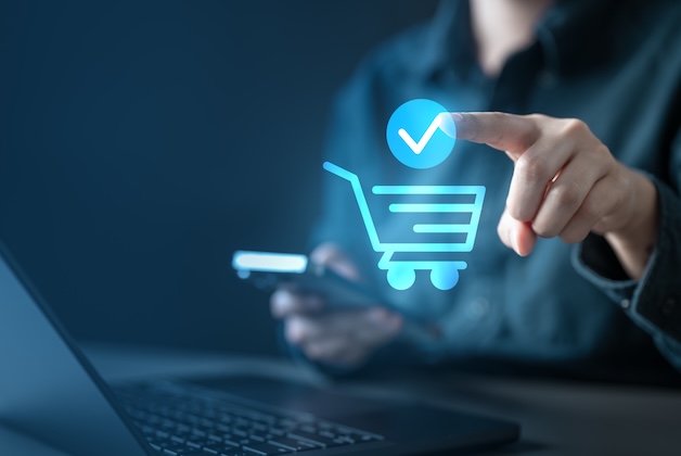 Mężczyzna dokonujący zakupu online za pomocą smartfona i laptopa – widoczna jest niebieska ikona koszyka.
