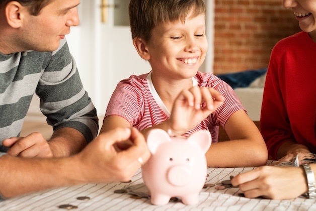 Uśmiechnięty chłopiec wrzuca monetę do różowej świnki skarbonki w obecności rodziców.