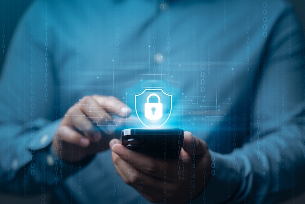 Mężczyzna trzymający smartfon, nad którym wyświetla się świecąca, niebieska ikona kłódki wpisanej w tarczę oraz pionowe ciągi cyfr binarnych, symbolizujące cyberbezpieczeństwo.