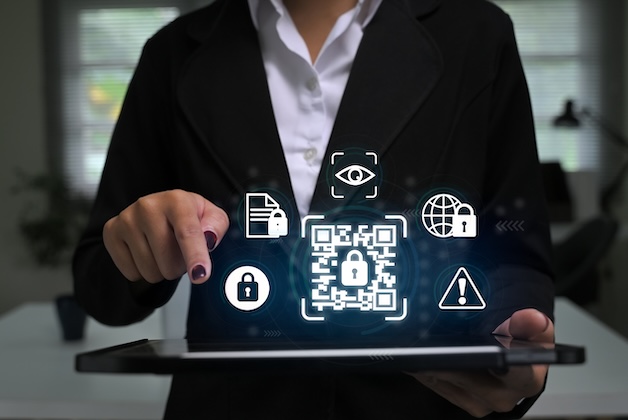 Kobieta w biznesowym garniturze trzyma tablet, nad którym unoszą się świecące ikony cyberbezpieczeństwa, w tym centralny kod QR z kłódką, symbol oka weryfikacji oraz trójkąt ostrzegawczy.