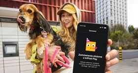 Uśmiechnięta dziewczyna trzymająca dużego psa na jednej ręce, w drugiej pokazująca smartfona z ikoną inpost. W tle widać paczkomat. 