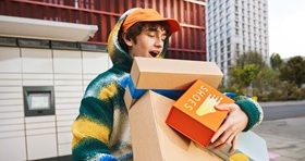 Młody, uśmiechnięty mężczyzna, trzyma w ramionach kilka pudełek po butach, idąc ulicą w mieście. Na jednym z pudełek widnieje napis "SHOES" oraz grafika stopy. W tle widać paczkomat.