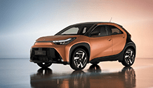 Miedziano-brązowa Toyota Aygo X Hybrid, zaparkowana w studiu z tłem w kolorach niebieskim i pomarańczowym. Auto stoi pod kątem, z widocznym przodem i lewym bokiem.