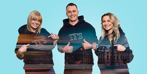 Trzy uśmiechnięte osoby w ciemnych bluzach z kapturem z napisem TFS, nałożone na obraz miasta nocą.