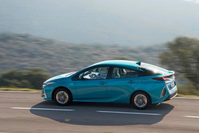 Zdjęcie przedstawia Toyotę Prius, w kolorze niebieskim, jadącą po asfaltowej drodze.  Samochód jest w ruchu, co sugeruje rozmyte tło, na którym widać porośnięte roślinnością doliny. 