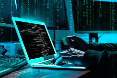 Ręce osoby pracującej przy laptopie, na ekranie kod komputerowy, w tle monitory – jedna dłoń trzyma kartę płatniczą; ilustracja zagrożeń związanych z bezpieczeństwem danych.