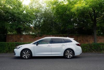 Biała Corolla TS Kombi zaparkowana przy ulicy, otoczona zielenią i drzewami, w tle kamienny mur i krzewy