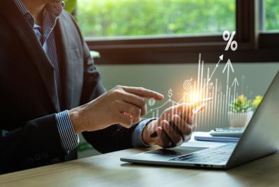 Biznesmen w garniturze korzysta ze smartfona przy biurku z laptopem; z telefonu emanują cyfrowe ikony i wykresy symbolizujące wzrost finansowy, procenty, dolary i strzałki w górę.