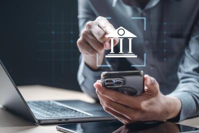 Mężczyzna w szarej koszuli używa smartfona, a na ekranie wyświetla się ikona banku. W tle widać laptopa.