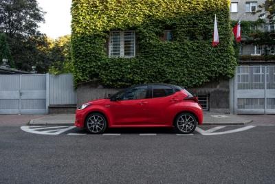 Czerwony hatchback - Toyota Yaris - zaparkowany na ulicy, na tle budynku całkowicie pokrytego bluszczem, z widoczną polską flagą.