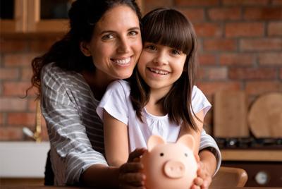 Zdjęcie przedstawia uśmiechniętą mamę i córkę, trzymające świnkę skarbonkę w kuchni. Jest to symboliczny obrazek związany z oszczędzaniem pieniędzy i edukacją finansową dzieci.