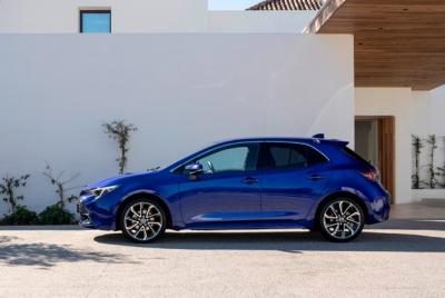 Zdjęcie przedstawia niebieską Toyotę Corollę Hatchback, widzianą z boku, zaparkowaną przed białym, nowoczesnym budynkiem.