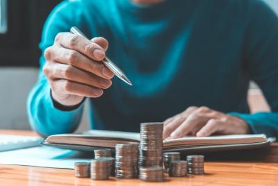 Zbliżenie na osobę w niebieskim swetrze trzymającą srebrny długopis i wskazującą nim na otwarty notes leżący za stosami monet w pierwszym planie. Scena symbolizuje planowanie budżetu, obliczanie oszczędności, inwestowanie lub analizę finansową.