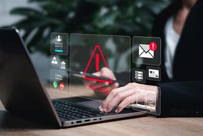 Zdjęcie przedstawia osobę pracującą na laptopie z nakładającymi się wirtualnymi ikonami alertów bezpieczeństwa, w tym czerwonym trójkątem ostrzegawczym i powiadomieniem e-mail.