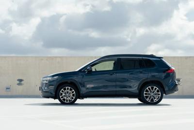 Zdjęcie przedstawia ciemnoniebieską Toyotę Corollę Cross 2025 zaparkowaną na zewnątrz na białym betonowym podłożu, z widokiem z boku.