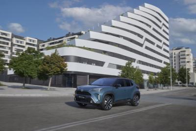 Niebieski samochód Toyota Yaris Cross jedzie ulicą miejską w słoneczny dzień. W tle znajduje się nowoczesny, biały budynek mieszkalny o falistej architekturze oraz inne bloki mieszkalne i drzewa.