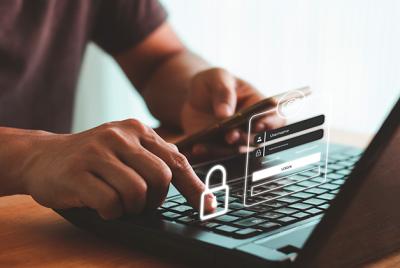 Zdjęcie przedstawia ręce osoby wpisującej dane na klawiaturze laptopa. Obok leży smartfon z ekranem logowania i symbolem kłódki, co sugeruje bezpieczeństwo online.