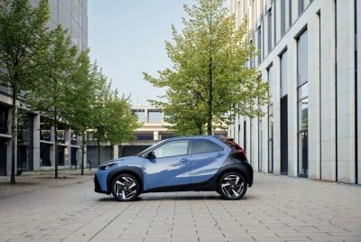 Niebieski samochód Toyota Aygo X zaparkowany na brukowanym chodniku w otoczeniu nowoczesnych budynków biurowych i drzew.
