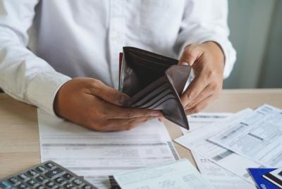 Zdjęcie przedstawia zbliżenie na ręce osoby w białej koszuli, która trzyma otwarty, pusty, czarny portfel nad stołem pokrytym licznymi rachunkami, dokumentami i kalkulatorem. Scena sugeruje problemy finansowe lub brak pieniędzy.