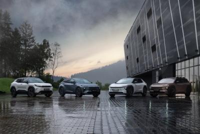 Cztery elektryczne SUV-y Toyota bZ4X Touring w różnych kolorach (od lewej: biały, szary, biały i brązowy) zaparkowane w rzędzie na mokrej, wybrukowanej powierzchni obok nowoczesnego budynku, z zielonymi wzgórzami i zachmurzonym niebem w tle.
