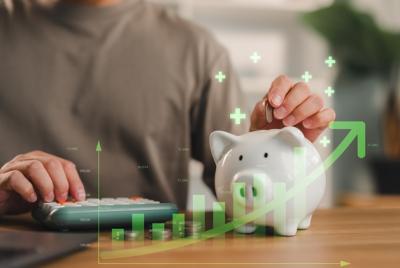 Osoba wkładająca srebrną monetę do białej skarbonki, trzymając drugą rękę na kalkulatorze. Na zdjęciu widoczne są nałożone cyfrowe wykresy wzrostu, zielone znaki plus i strzałka w górę, symbolizujące oszczędzanie, inwestowanie i wzrost finansowy.