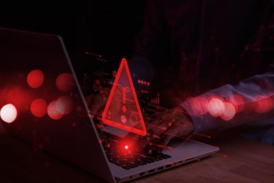 Zbliżenie dłoni osoby pracującej na laptopie w ciemnym otoczeniu, z nałożonym świecącym czerwonym trójkątem ostrzegawczym (ikona zagrożenia cybernetycznego) i rozmytymi czerwonymi światłami w tle, ilustrujące naruszenie bezpieczeństwa.