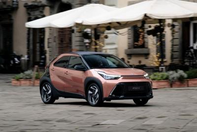 Zdjęcie przedstawia nowoczesny, miedziano-czarny miejski crossover Toyota Aygo X jadący po brukowanym placu na tle kawiarnianych parasoli i zabytkowych kamienic. 