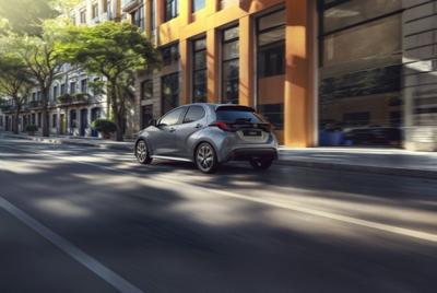 Zdjęcie na którym widoczna jest srebrna Toyota Yaris w ruchu, na tle nowoczesnej miejskiej architektury i drzew, widok od tyłu.